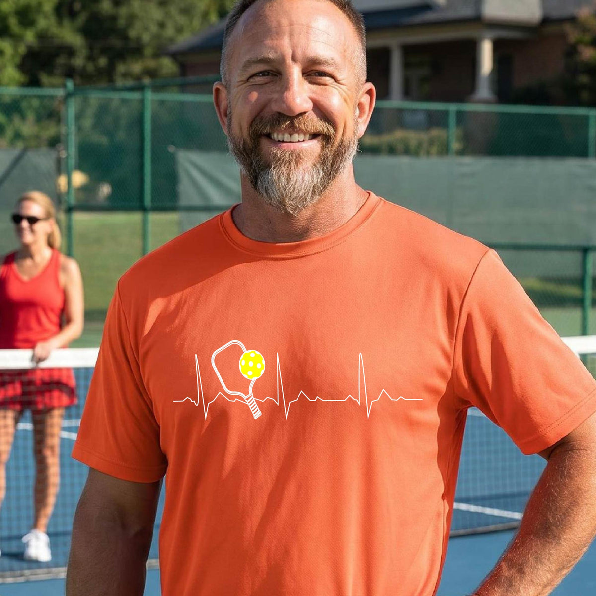 A close-up of a man in an orange short-sleeve t-shirt sporting a white pickleball graphic of a heartbeat EKG including a pickleball paddle and a yellow pickleball across the check, in front of a pickleball court.