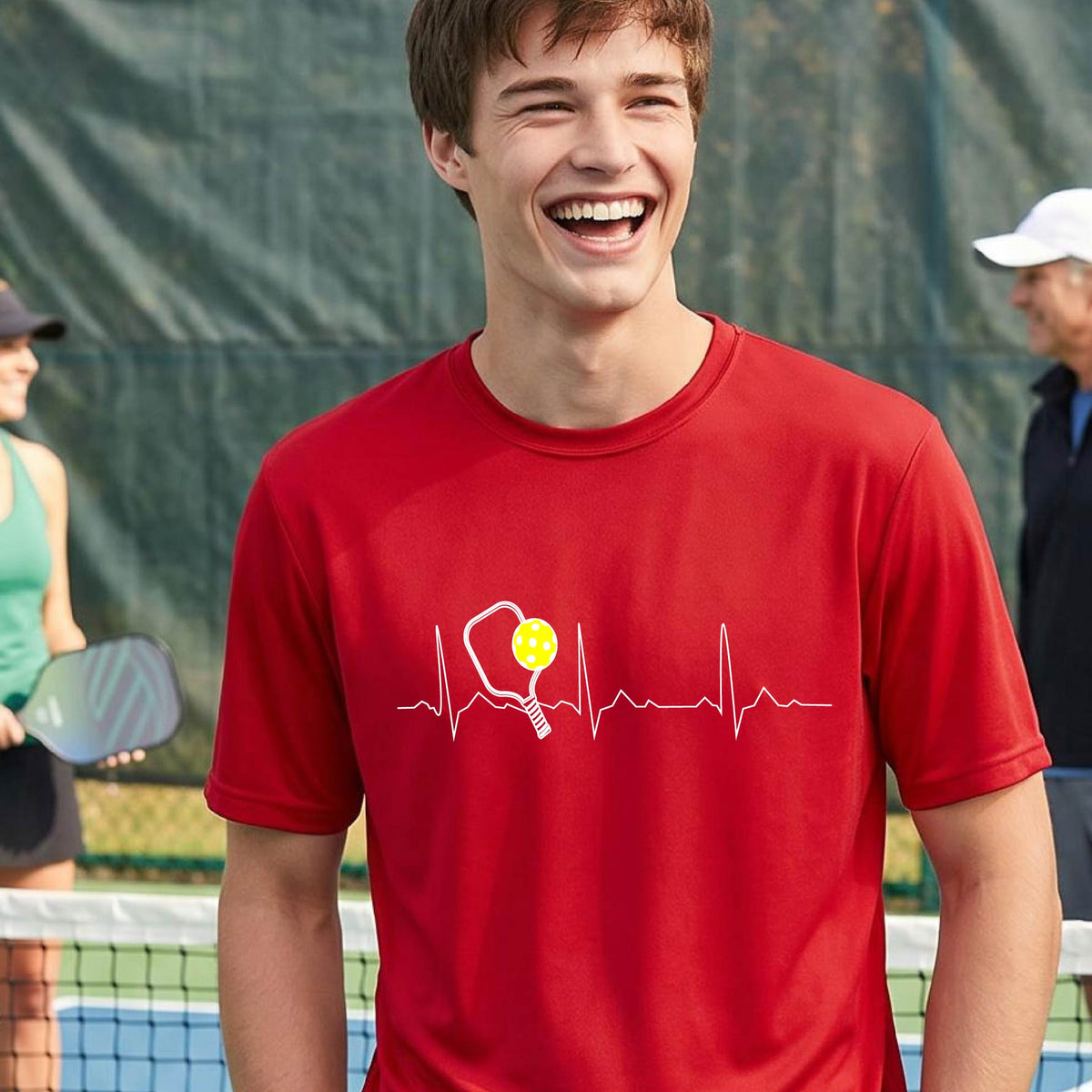 A close-up of a man in a red short-sleeve t-shirt sporting a white pickleball graphic of a heartbeat EKG including a pickleball paddle and a yellow pickleball across the check, in front of a pickleball court.