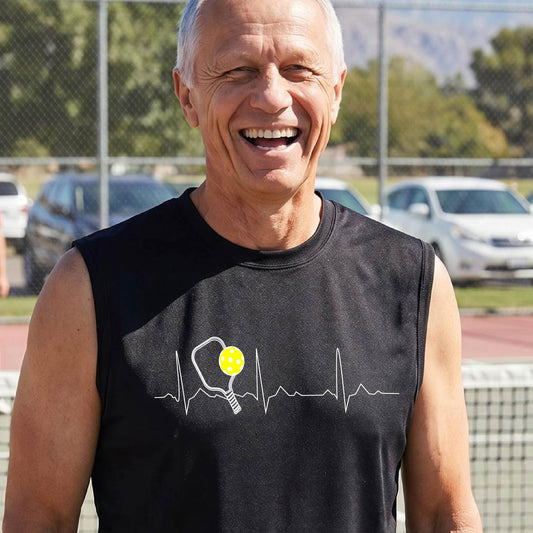 A close-up of a man in a black sleeveless t-shirt sporting a white pickleball graphic of a heartbeat EKG including a pickleball paddle and a yellow pickleball across the check, in front of a pickleball court.