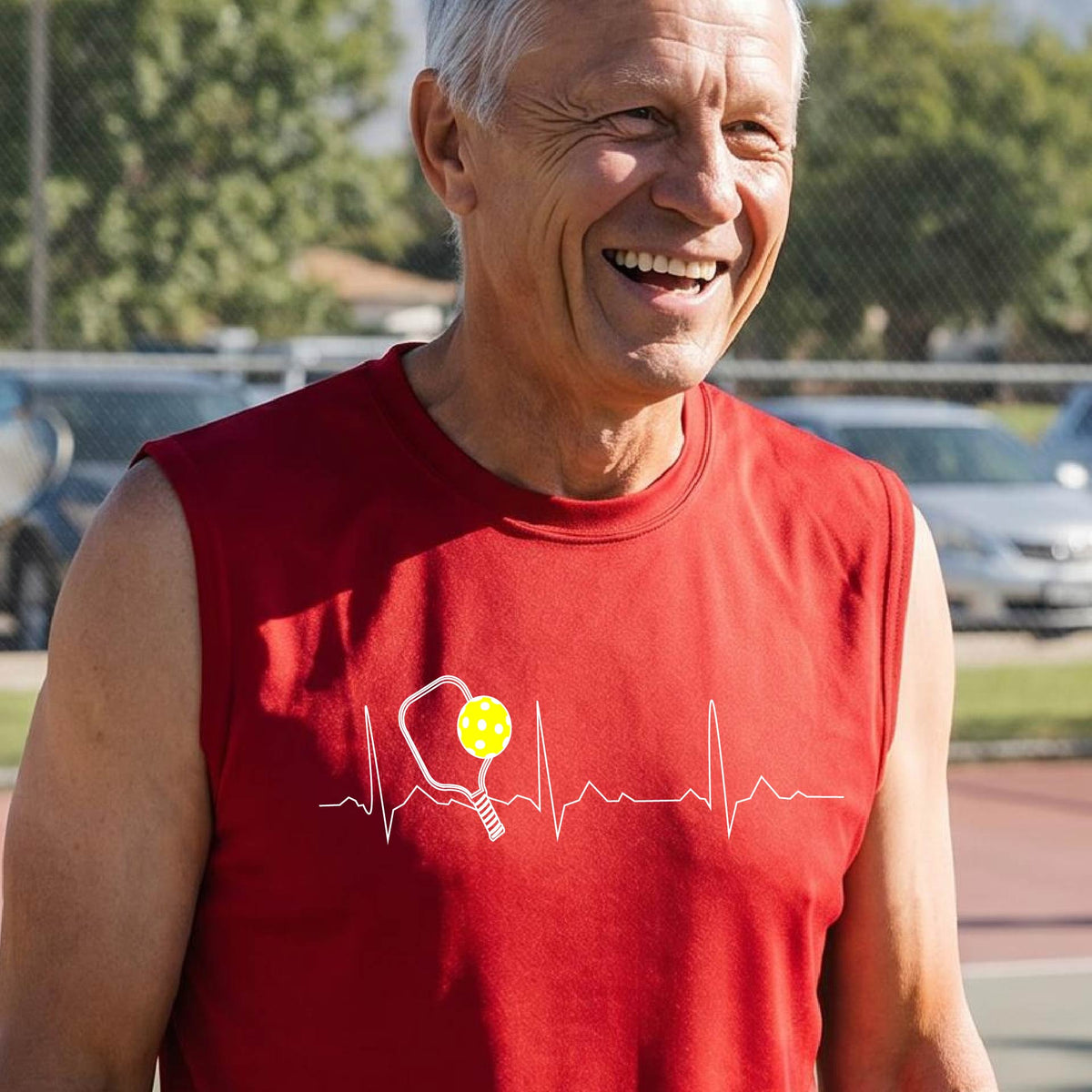 A close-up of a man in a red sleeveless t-shirt sporting a white pickleball graphic of a heartbeat EKG including a pickleball paddle and a yellow pickleball across the check, in front of a pickleball court.