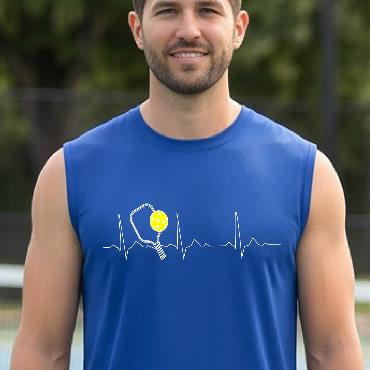 A close-up of a man in a royal blue sleeveless t-shirt sporting a white pickleball graphic of a heartbeat EKG including a pickleball paddle and a yellow pickleball across the check, in front of a pickleball court.