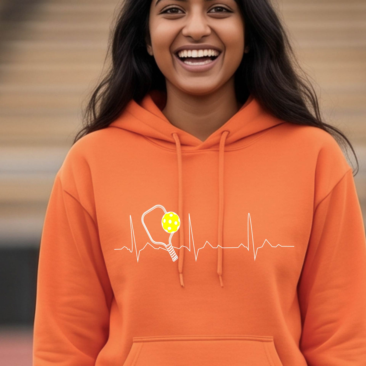 A close-up of a unisex orange hoodie pullover showing a white pickleball graphic of a heartbeat EKG including a pickleball paddle and a yellow pickleball, in front of a pickleball court.