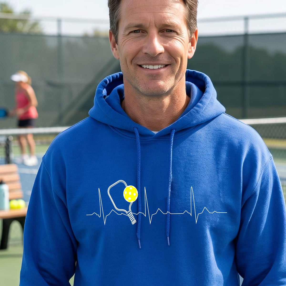 A close-up of a unisex royal blue hoodie pullover showing a white pickleball graphic of a heartbeat EKG including a pickleball paddle and a yellow pickleball, in front of a pickleball court.