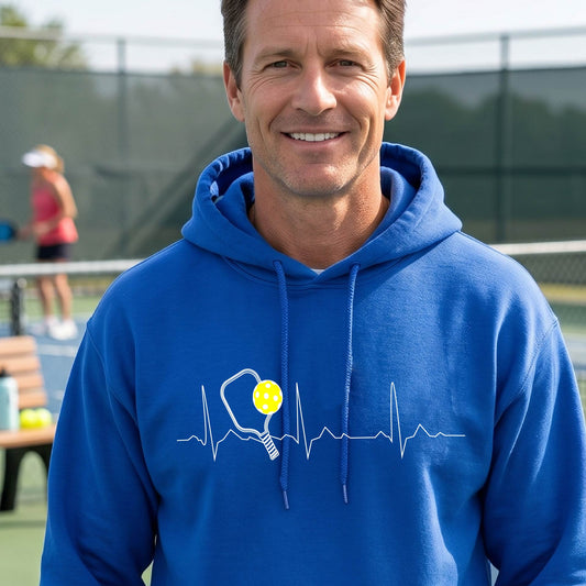 A close-up of a unisex royal blue hoodie pullover showing a white pickleball graphic of a heartbeat EKG including a pickleball paddle and a yellow pickleball, in front of a pickleball court.