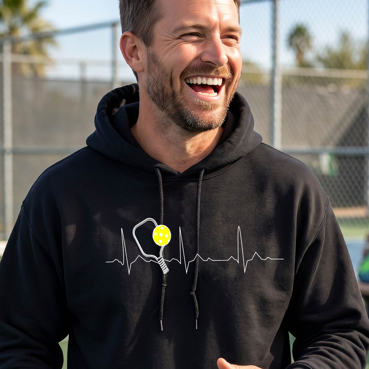 A close-up of a unisex black hoodie pullover showing a white pickleball graphic of a heartbeat EKG including a pickleball paddle and a yellow pickleball, in front of a pickleball court.