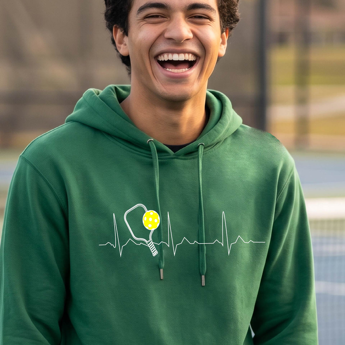 A close-up of a unisex green hoodie pullover showing a white pickleball graphic of a heartbeat EKG including a pickleball paddle and a yellow pickleball, in front of a pickleball court.