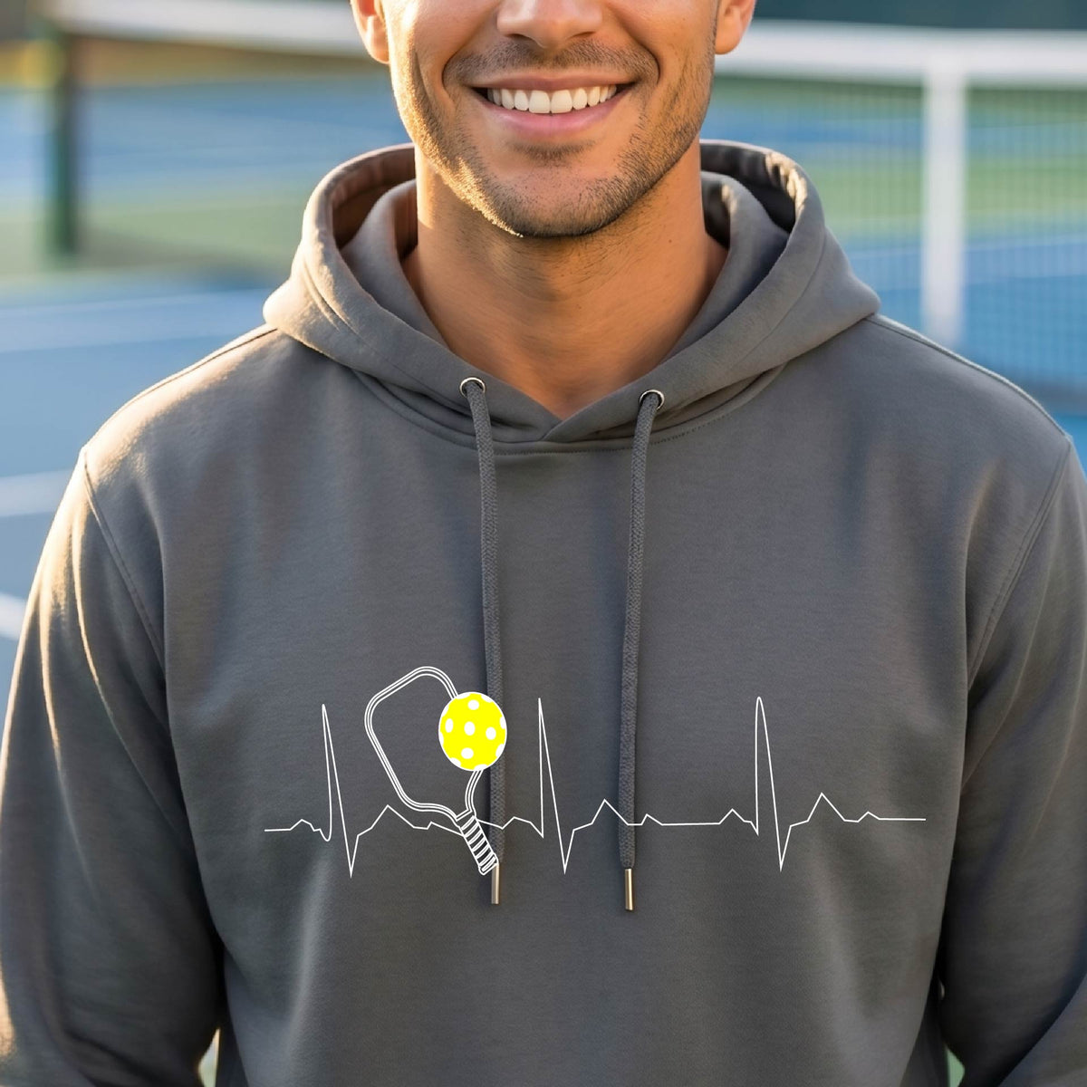 A close-up of a unisex dark gray hoodie pullover showing a white pickleball graphic of a heartbeat EKG including a pickleball paddle and a yellow pickleball, in front of a pickleball court.