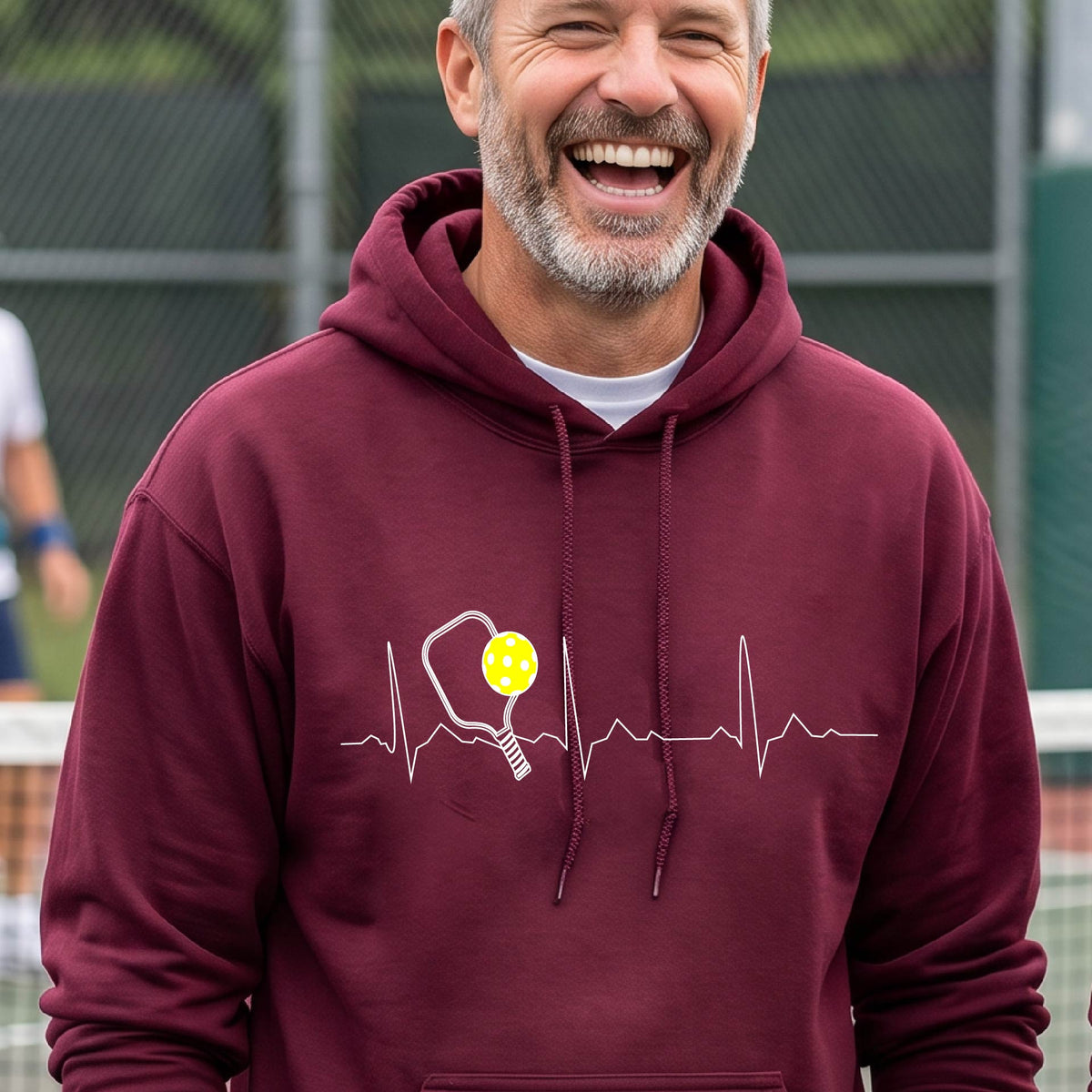 A close-up of a unisex rmaroon hoodie pullover showing a white pickleball graphic of a heartbeat EKG including a pickleball paddle and a yellow pickleball, in front of a pickleball court.