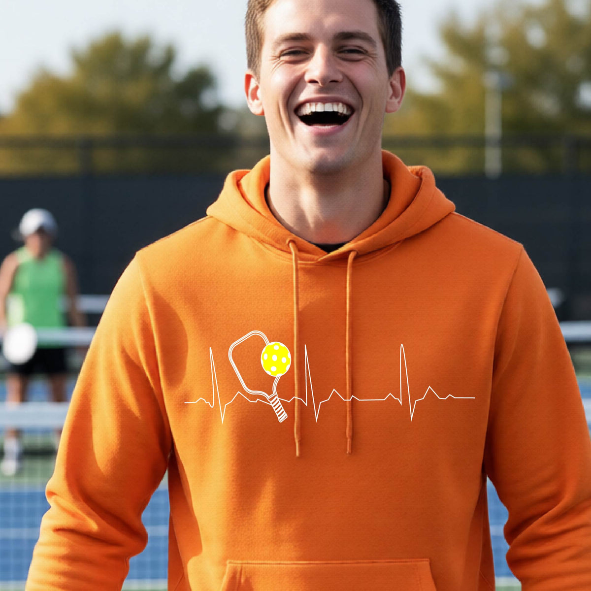 A close-up of a unisex orange hoodie pullover showing a white pickleball graphic of a heartbeat EKG including a pickleball paddle and a yellow pickleball, in front of a pickleball court.