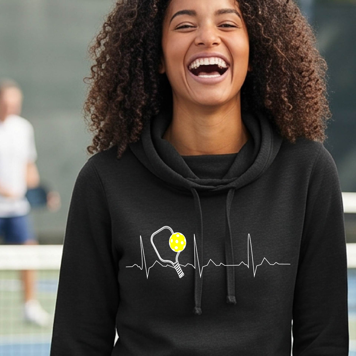 A close up of a woman in a black cowl-neck hoodie pullover showing a white pickleball graphic of a heartbeat EKG including a pickleball paddle and a yellow pickleball, in front of a pickleball court.