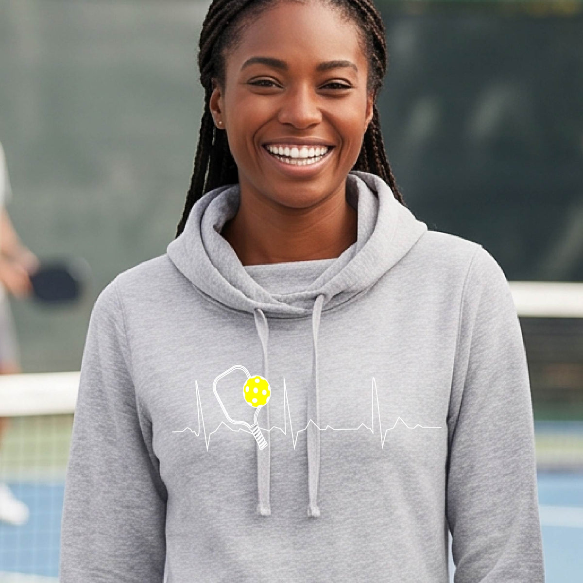 A close up of a woman in a light gray cowl-neck hoodie pullover showing a white pickleball graphic of a heartbeat EKG including a pickleball paddle and a yellow pickleball, in front of a pickleball court.