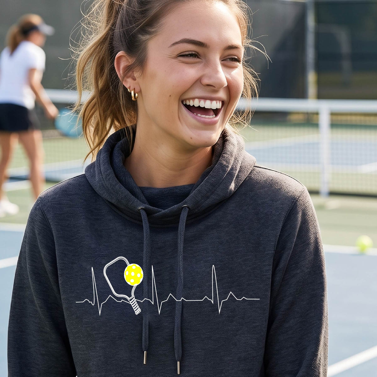 A close up of a woman in a navy blue cowl-neck hoodie pullover showing a white pickleball graphic of a heartbeat EKG including a pickleball paddle and a yellow pickleball, in front of a pickleball court.