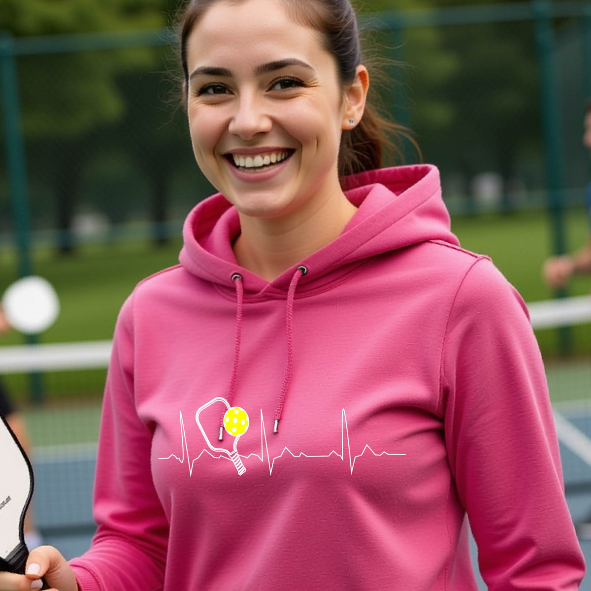 A close up of a woman in a pink cowl-neck hoodie pullover showing a white pickleball graphic of a heartbeat EKG including a pickleball paddle and a yellow pickleball, in front of a pickleball court.