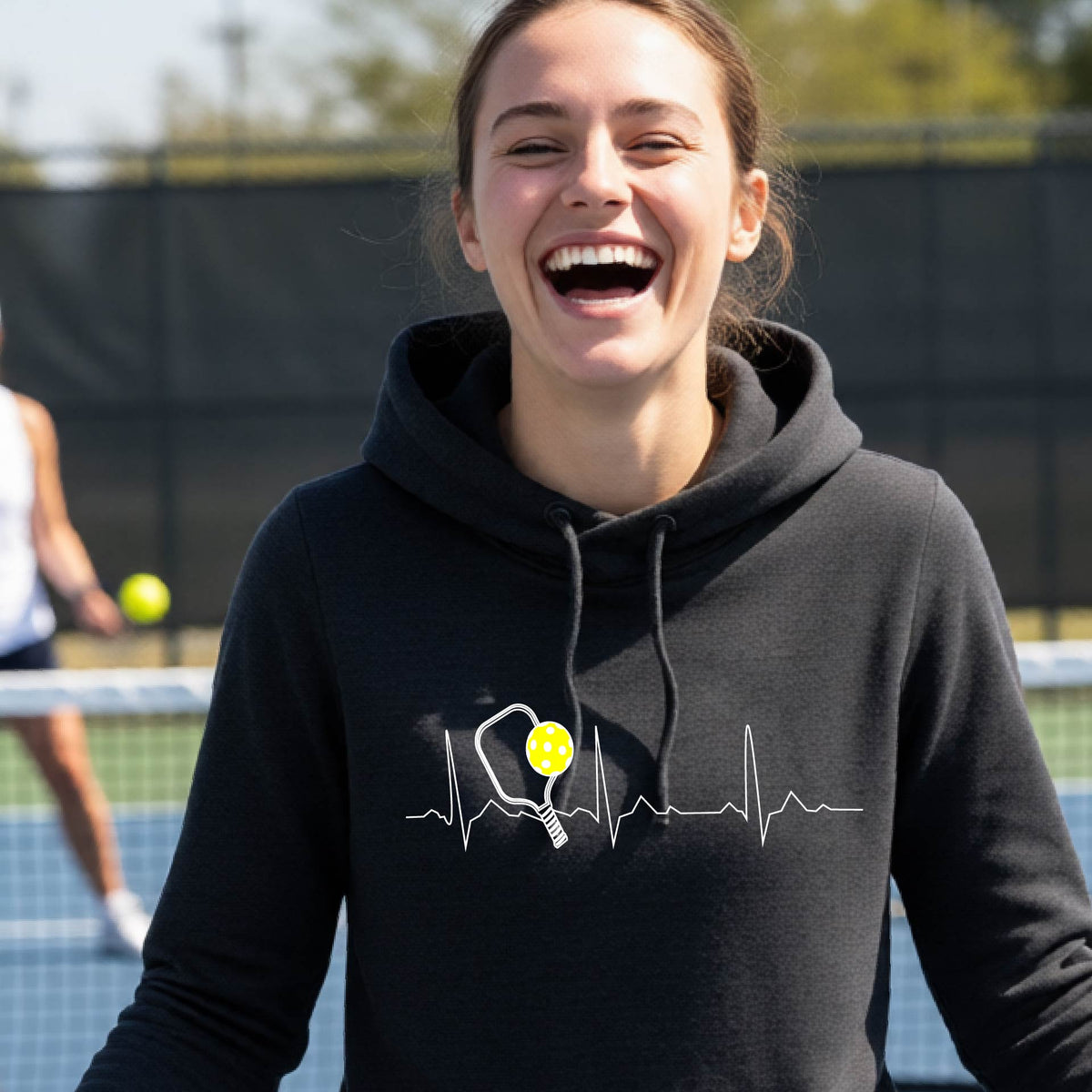 A close-up of a woman wearing a fitted black hoodie pullover showing a white pickleball graphic of a heartbeat EKG including a pickleball paddle and a yellow pickleball, in front of a pickleball cour