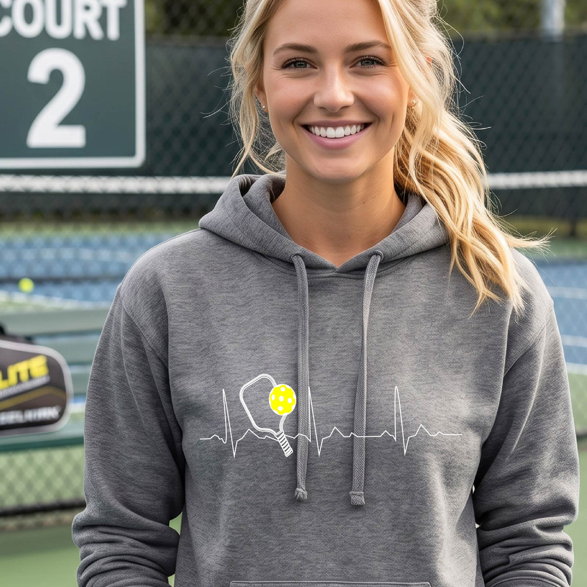 A close-up of a woman wearing a fitted gray hoodie pullover showing a white pickleball graphic of a heartbeat EKG including a pickleball paddle and a yellow pickleball, in front of a pickleball court.