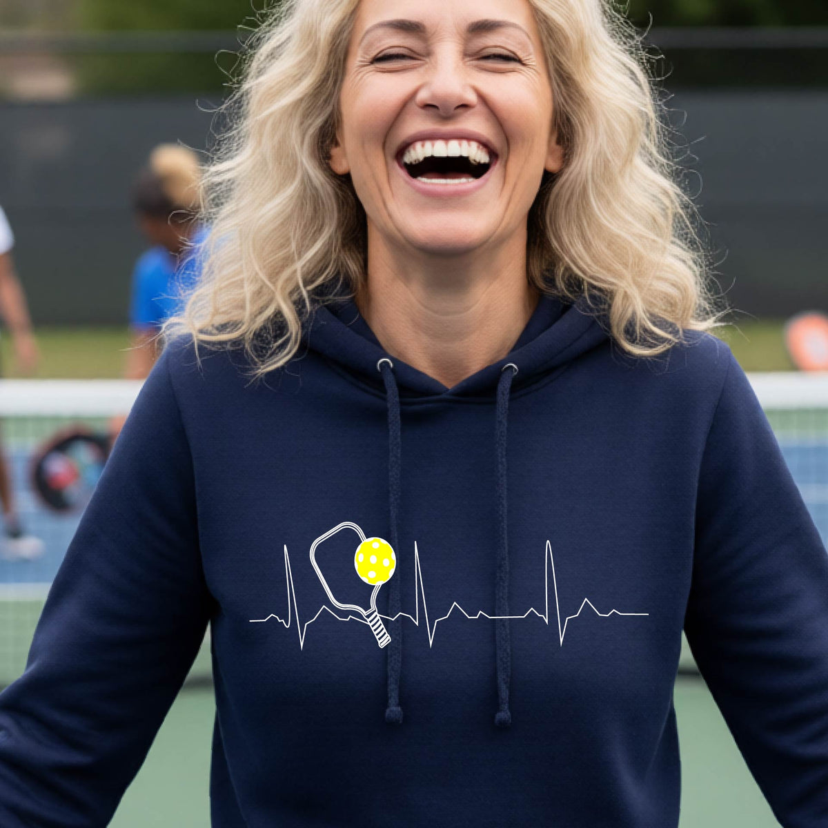 A close-up of a woman wearing a fitted navy blue hoodie pullover showing a white pickleball graphic of a heartbeat EKG including a pickleball paddle and a yellow pickleball, in front of a pickleball cour