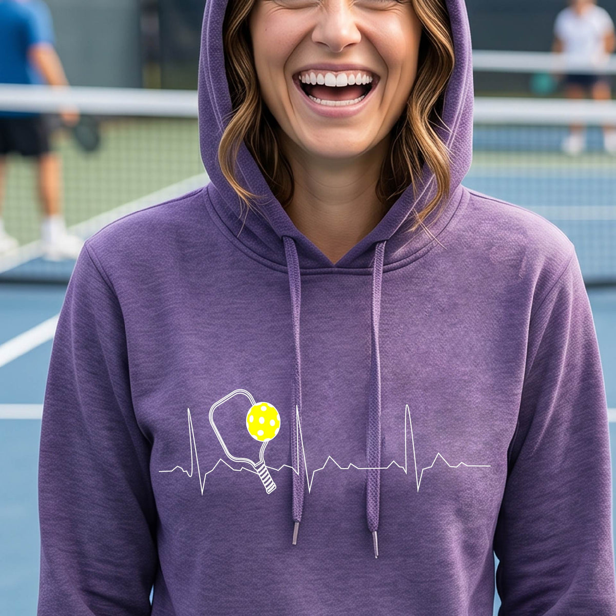 A close-up of a woman wearing a fitted purple hoodie pullover showing a white pickleball graphic of a heartbeat EKG including a pickleball paddle and a yellow pickleball, in front of a pickleball cour