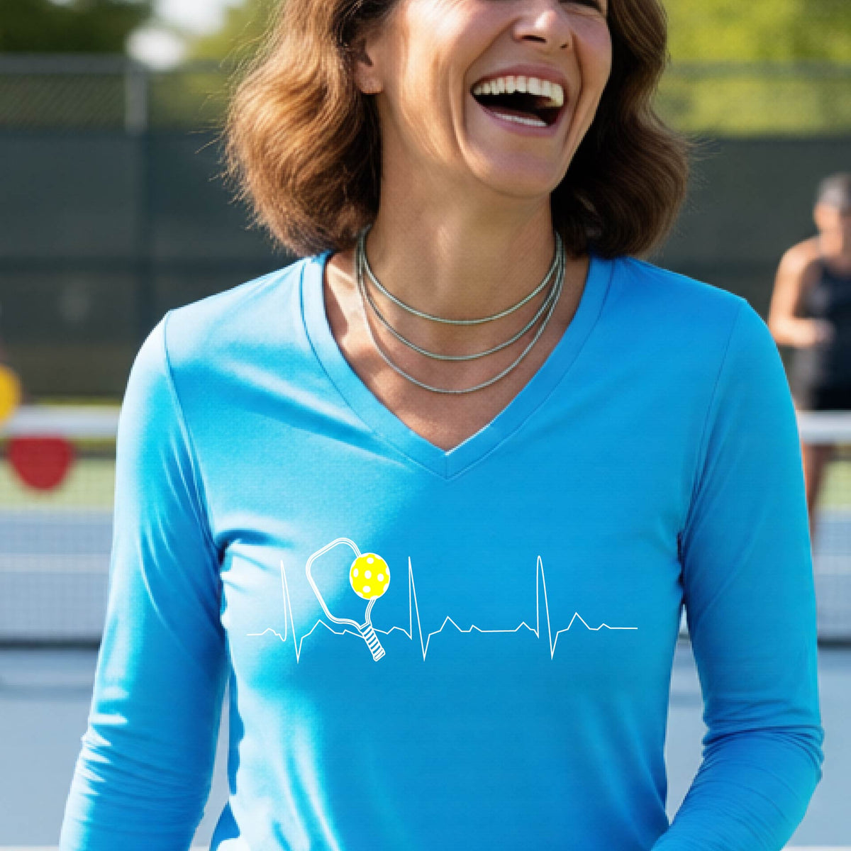 A close-up of a womens in a bright blue long-sleeve t-shirt sporting a white pickleball graphic of a heartbeat EKG including a pickleball paddle and a yellow pickleball across the check, in front of a pickleball court.
