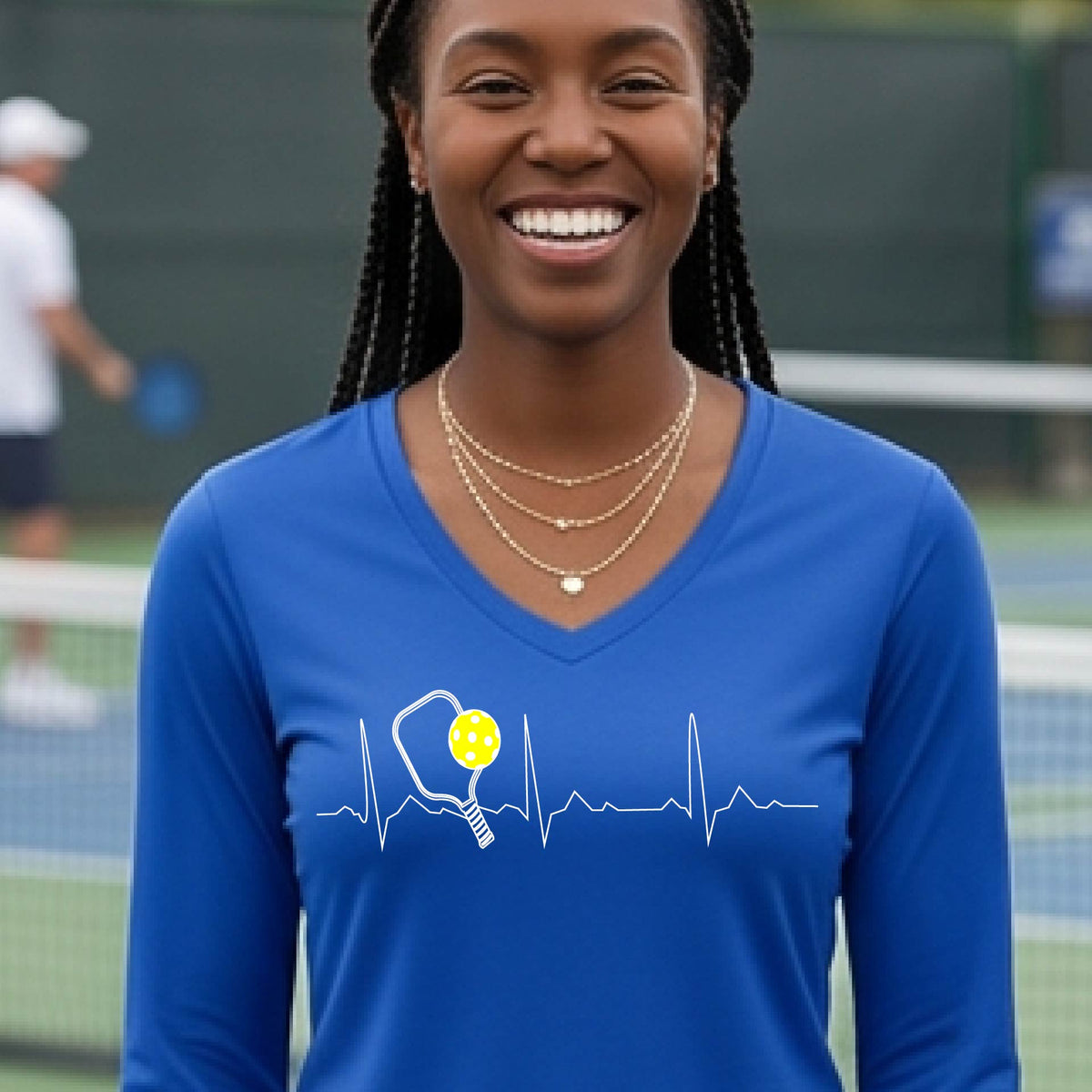 A close-up of a womens in a royal blue long-sleeve t-shirt sporting a white pickleball graphic of a heartbeat EKG including a pickleball paddle and a yellow pickleball across the check, in front of a pickleball court.
