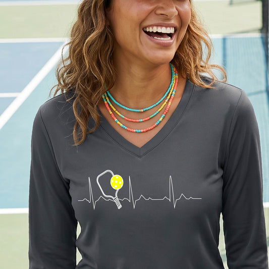 A close-up of a womens in a gray long-sleeve t-shirt sporting a white pickleball graphic of a heartbeat EKG including a pickleball paddle and a yellow pickleball across the check, in front of a pickleball court.

