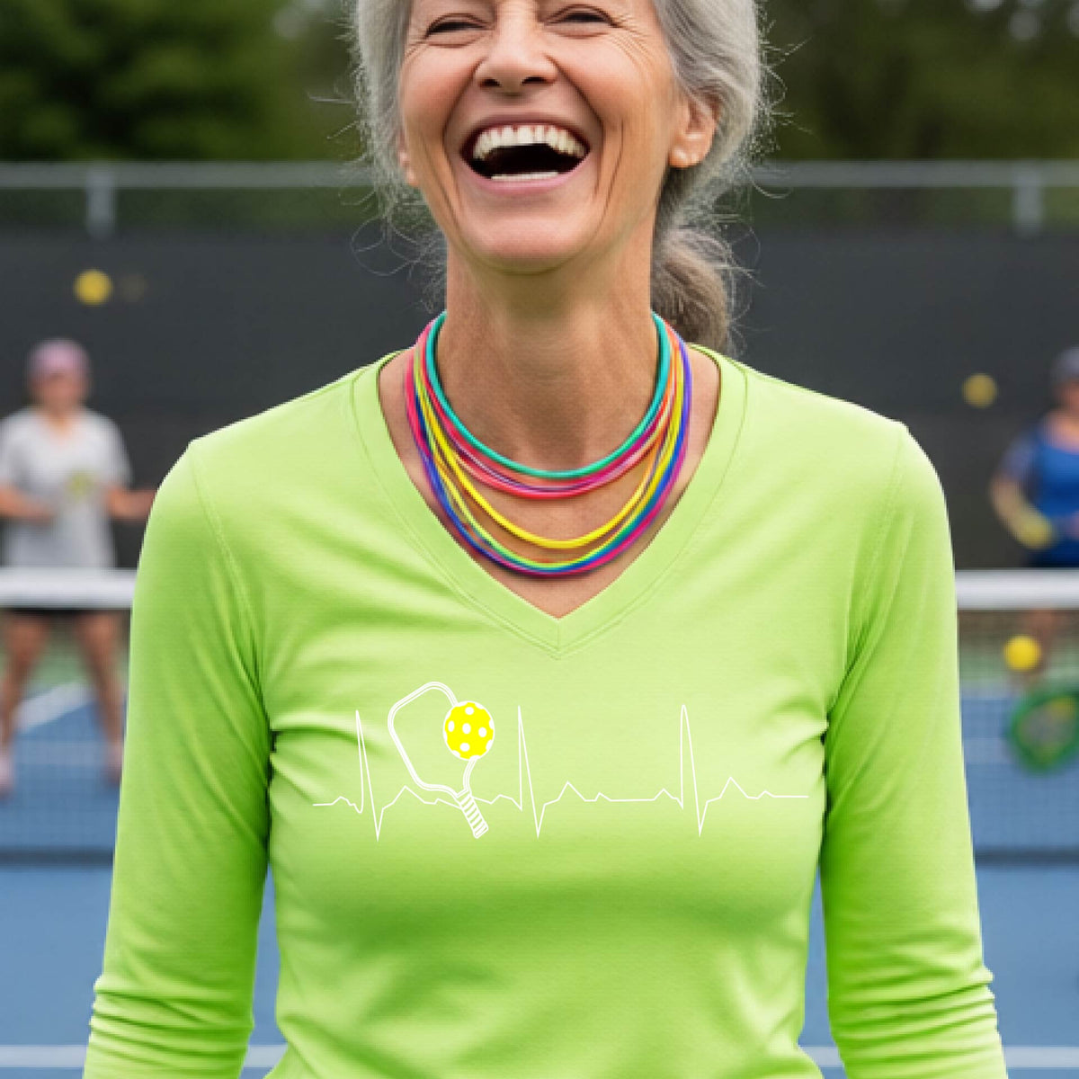 A close-up of a womens in a lime green long-sleeve t-shirt sporting a white pickleball graphic of a heartbeat EKG including a pickleball paddle and a yellow pickleball across the check, in front of a pickleball court.

