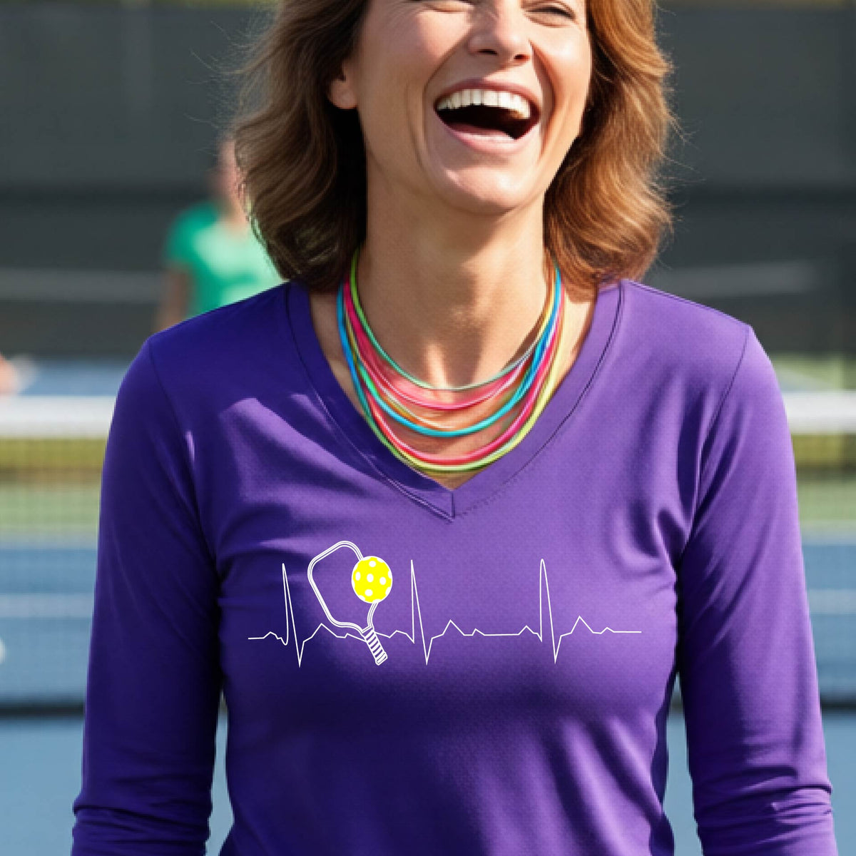 A close-up of a womens in a  long-sleeve t-shirt sporting a white pickleball graphic of a heartbeat EKG including a pickleball paddle and a yellow pickleball across the check, in front of a pickleball court.
urple