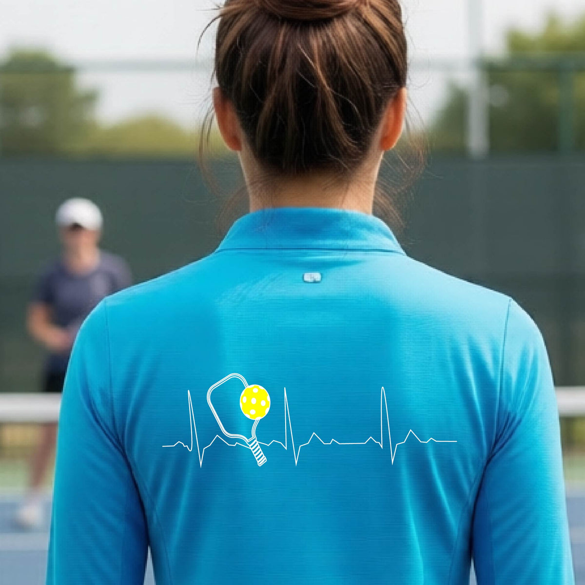 A close-up of a woman wearing a bright blue quarter-zip shirt with  a white pickleball graphic of a heartbeat EKG including a pickleball paddle and a yellow pickleball on the back, in front of a pickleball court.