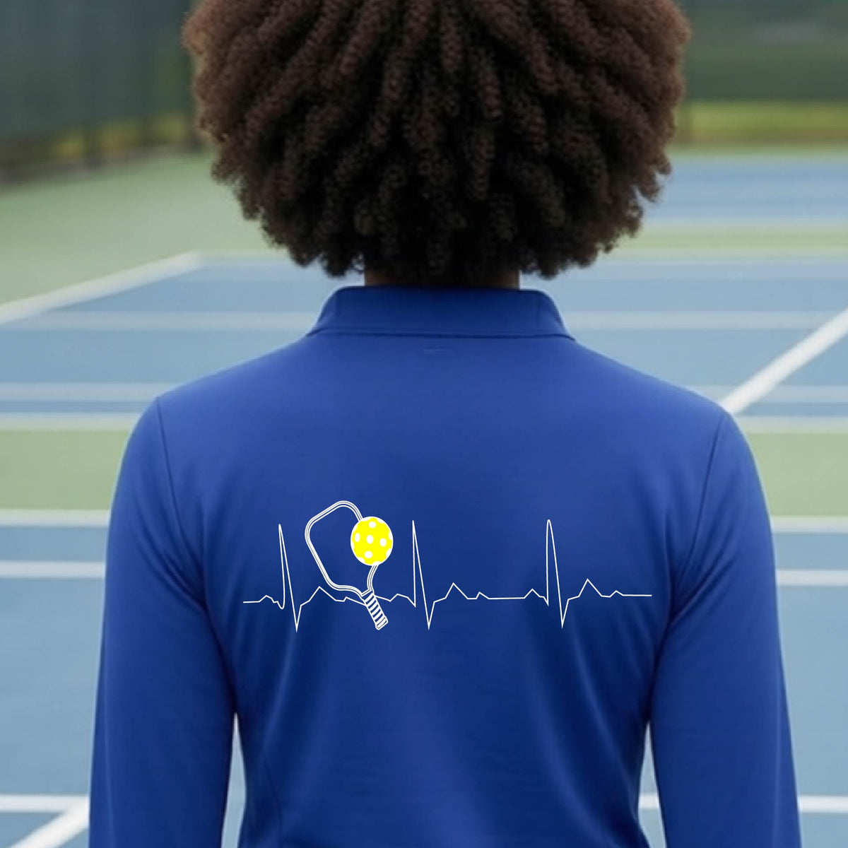 A close-up of a royal blue womens long-sleeve quarter-zip shirt showing a white pickleball graphic of a heartbeat EKG including a pickleball paddle and a yellow pickleball, in front of a pickleball court.
