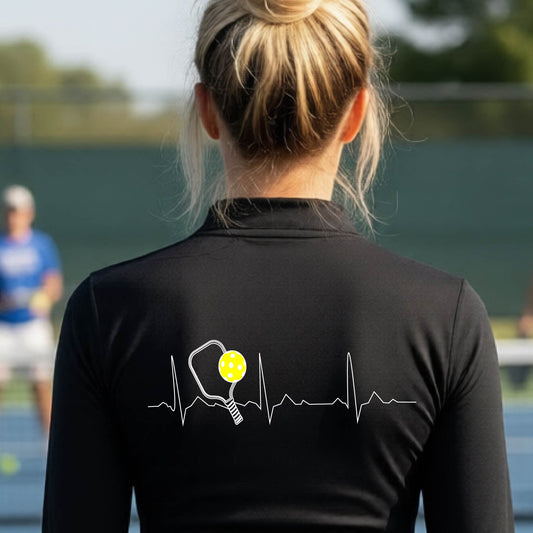 A close-up of a woman wearing a black quarter-zip shirt with  a white pickleball graphic of a heartbeat EKG including a pickleball paddle and a yellow pickleball on the back, in front of a pickleball court.