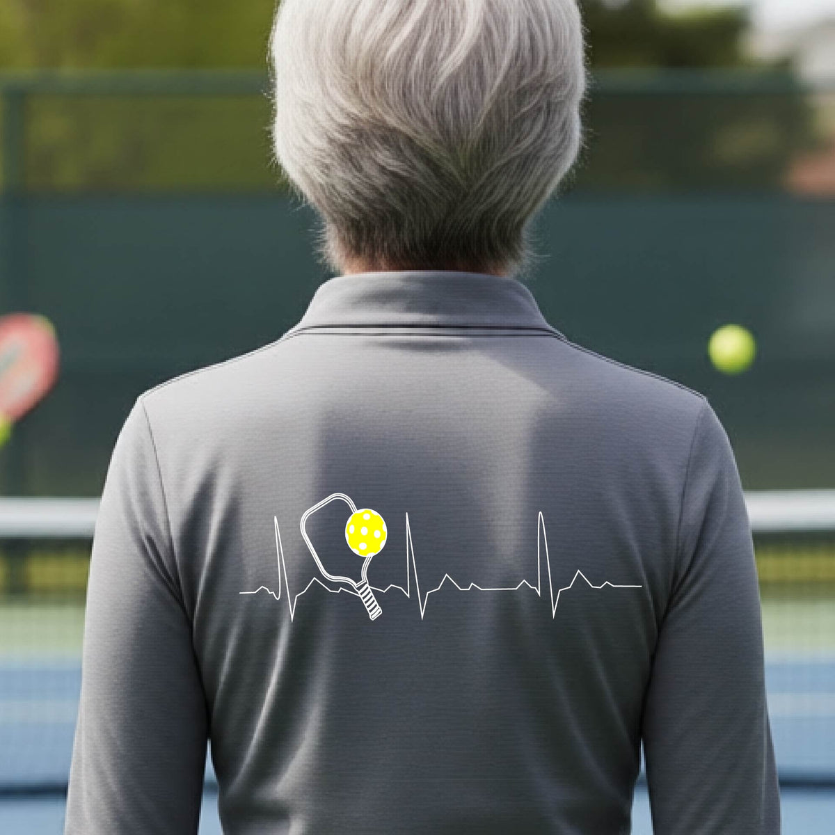 A close-up of a gray womens long-sleeve quarter-zip shirt showing a white pickleball graphic of a heartbeat EKG including a pickleball paddle and a yellow pickleball, in front of a pickleball court.