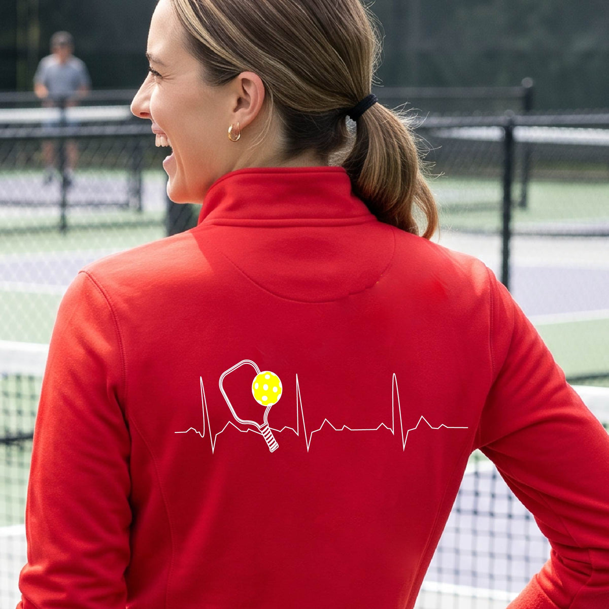 A close-up of a woman wearing a red quarter-zip shirt with  a white pickleball graphic of a heartbeat EKG including a pickleball paddle and a yellow pickleball on the back, in front of a pickleball court.