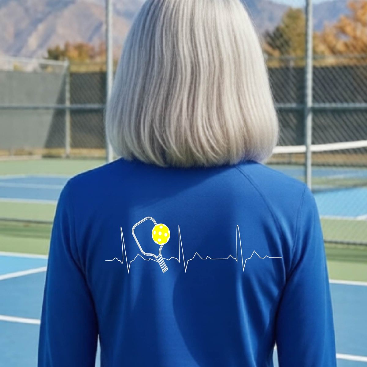A close-up of a woman wearing a royal blue quarter-zip shirt with  a white pickleball graphic of a heartbeat EKG including a pickleball paddle and a yellow pickleball on the back, in front of a pickleball court.