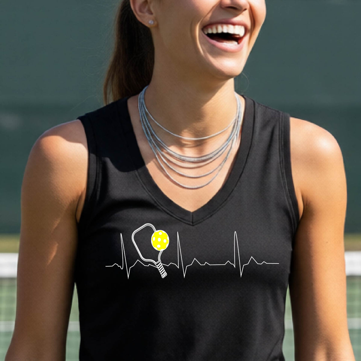 A close-up of a black womens racerback tank top showing a white pickleball graphic of a heartbeat EKG including a pickleball paddle and a yellow pickleball, in front of a pickleball court.