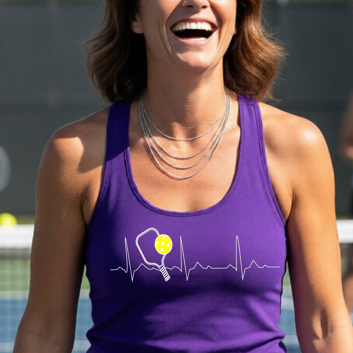 A close-up of a purple womens racerback tank top showing a white pickleball graphic of a heartbeat EKG including a pickleball paddle and a yellow pickleball, in front of a pickleball court.