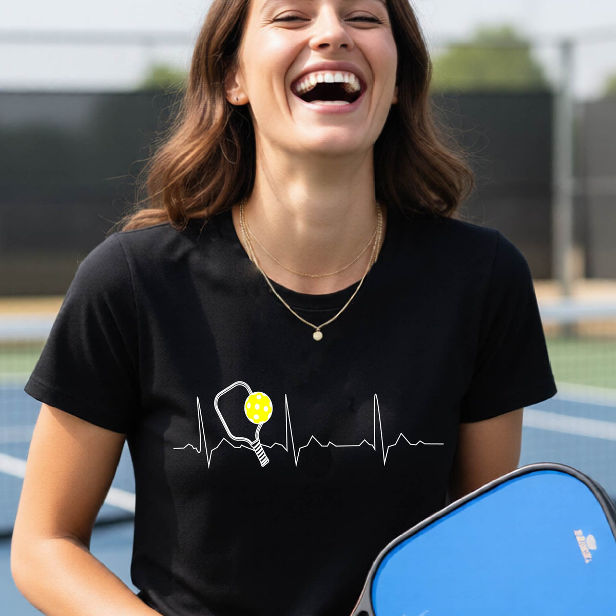 A close-up of a womens in a black short-sleeve t-shirt sporting a white pickleball graphic of a heartbeat EKG including a pickleball paddle and a yellow pickleball across the check, in front of a pickleball court.