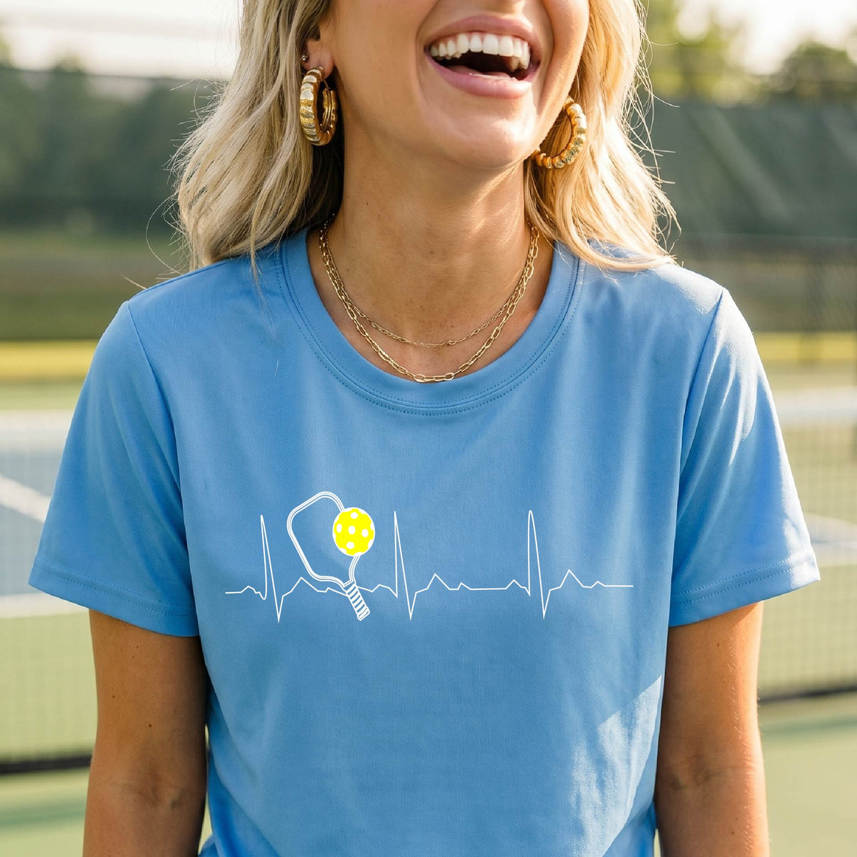 A close-up of a womens in a blue short-sleeve t-shirt sporting a white pickleball graphic of a heartbeat EKG including a pickleball paddle and a yellow pickleball across the check, in front of a pickleball court.