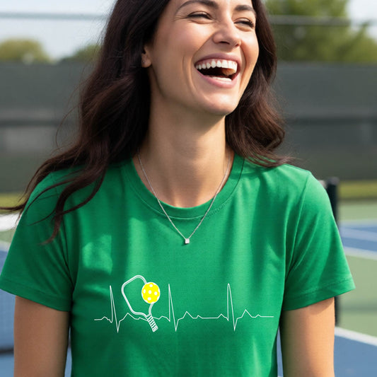 A close-up of a womens in a green short-sleeve t-shirt sporting a white pickleball graphic of a heartbeat EKG including a pickleball paddle and a yellow pickleball across the check, in front of a pickleball court.