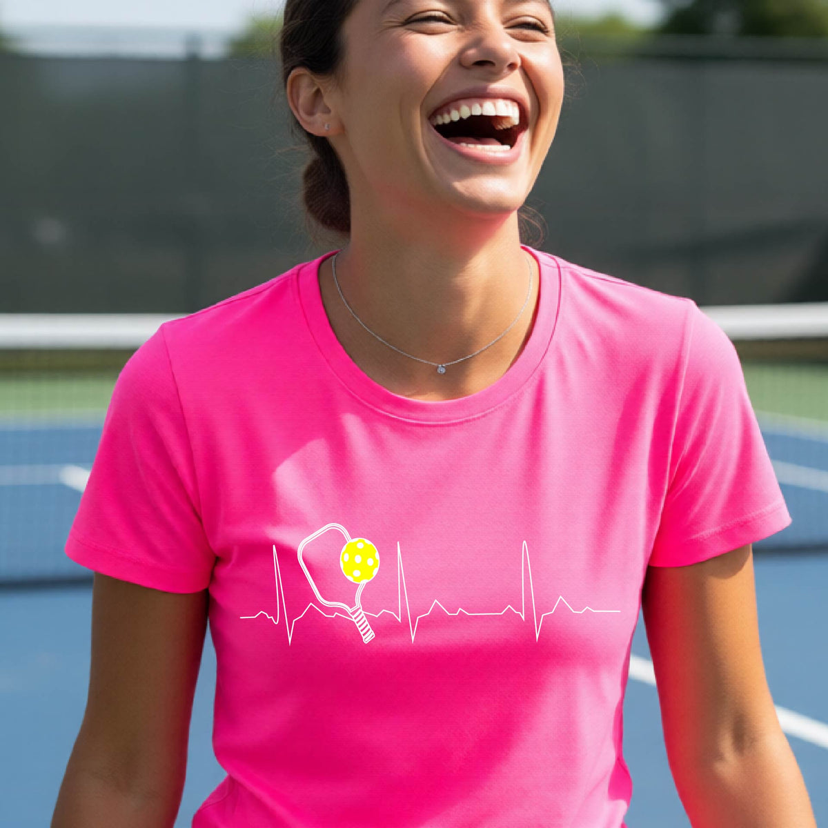 A close-up of a womens in a pink short-sleeve t-shirt sporting a white pickleball graphic of a heartbeat EKG including a pickleball paddle and a yellow pickleball across the check, in front of a pickleball court.