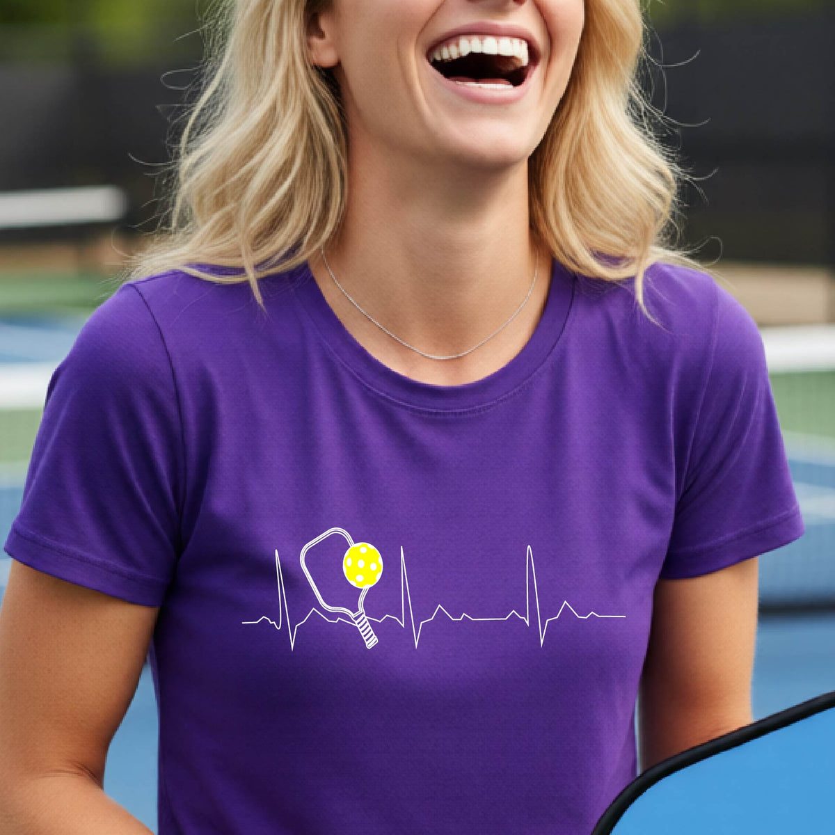 A close-up of a womens in a purple short-sleeve t-shirt sporting a white pickleball graphic of a heartbeat EKG including a pickleball paddle and a yellow pickleball across the check, in front of a pickleball court.