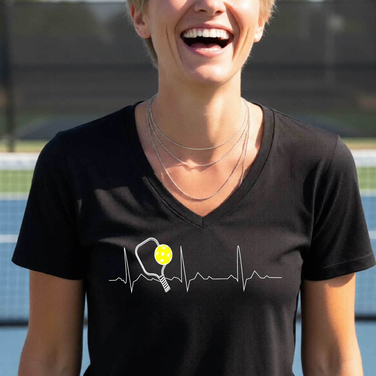A close-up of a womens in a black short-sleeve v-neck t-shirt sporting a white pickleball graphic of a heartbeat EKG including a pickleball paddle and a yellow pickleball across the check, in front of a pickleball court.
