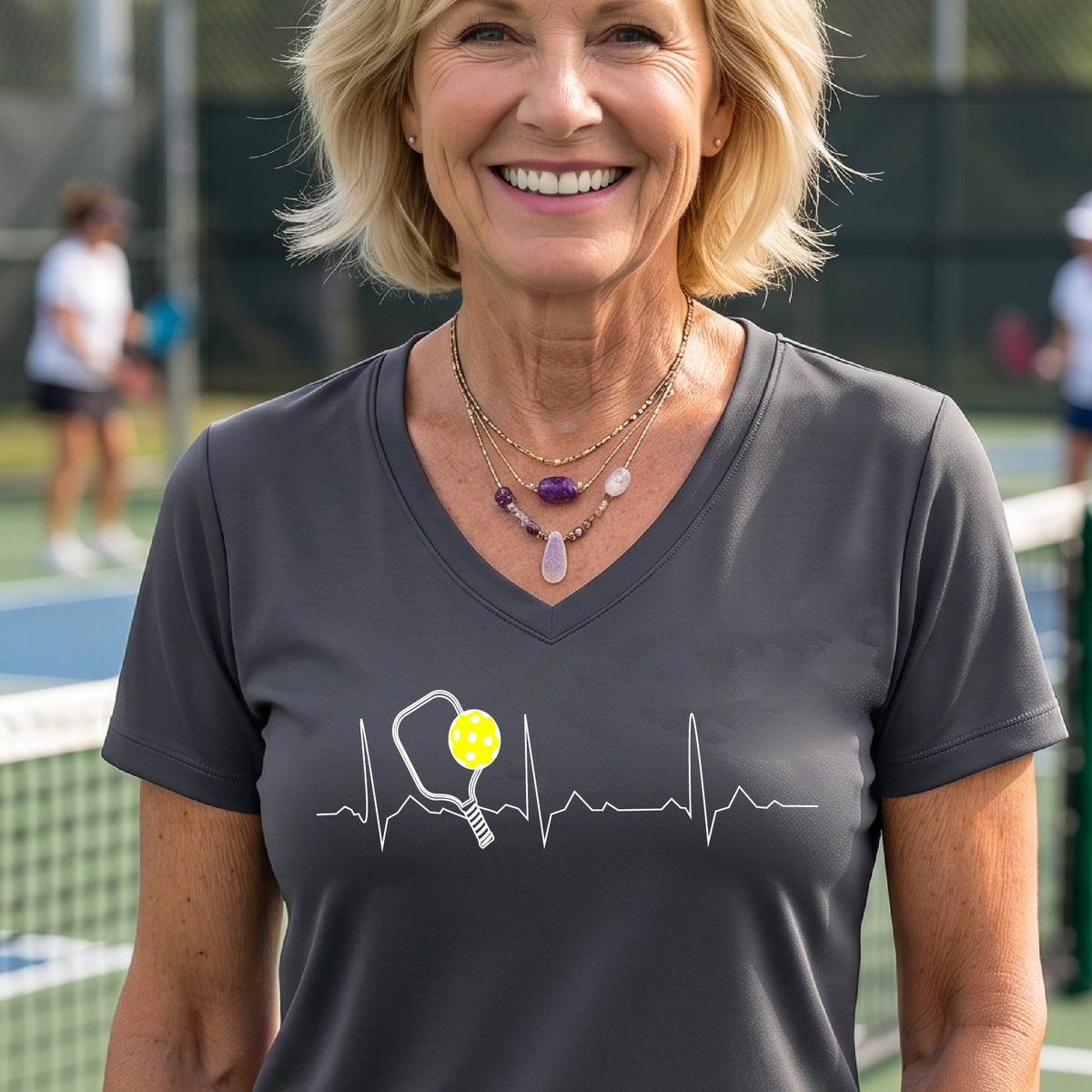 A close-up of a womens in a gray short-sleeve v-neck t-shirt sporting a white pickleball graphic of a heartbeat EKG including a pickleball paddle and a yellow pickleball across the check, in front of a pickleball court.