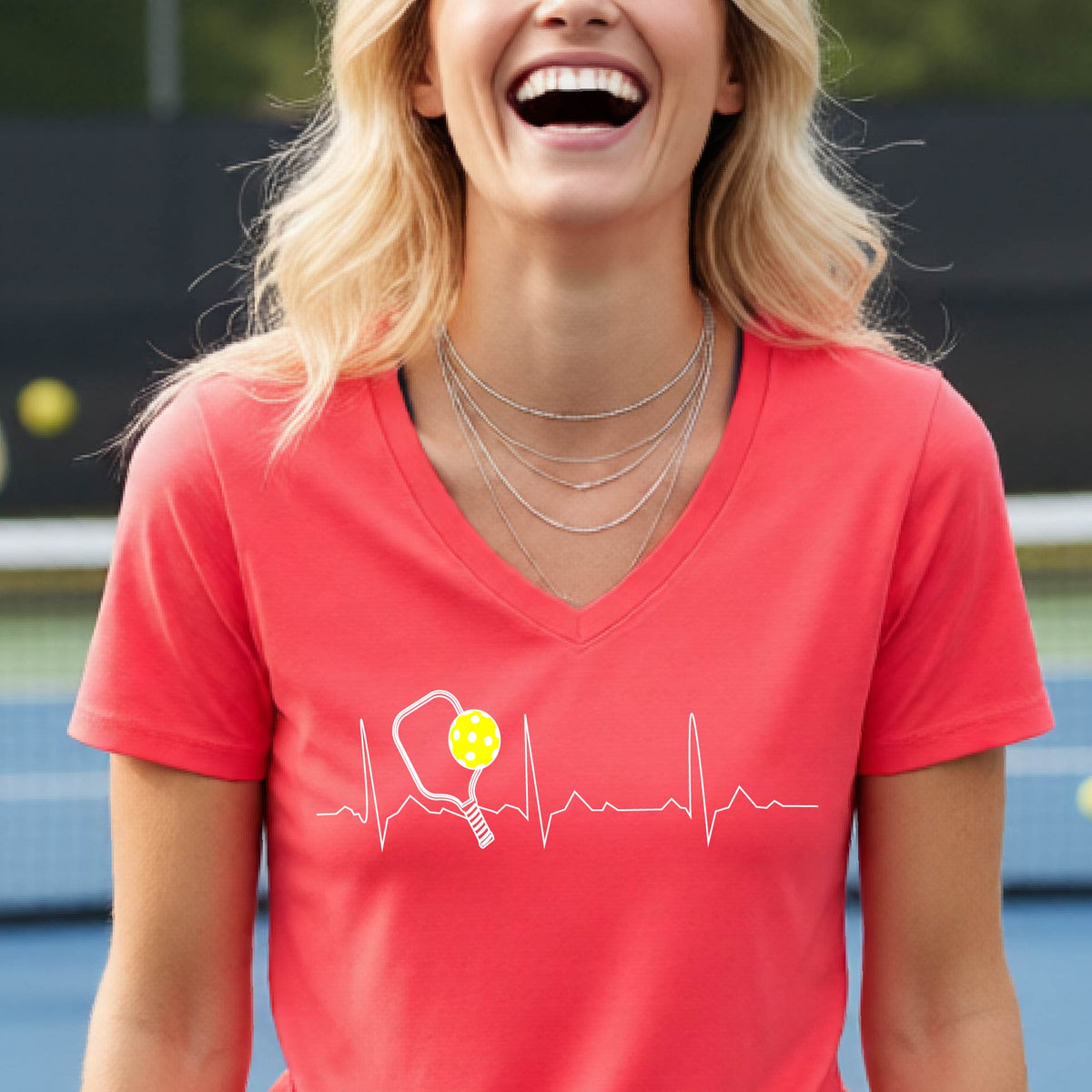 A close-up of a womens in a coral short-sleeve v-neck t-shirt sporting a white pickleball graphic of a heartbeat EKG including a pickleball paddle and a yellow pickleball across the check, in front of a pickleball court.