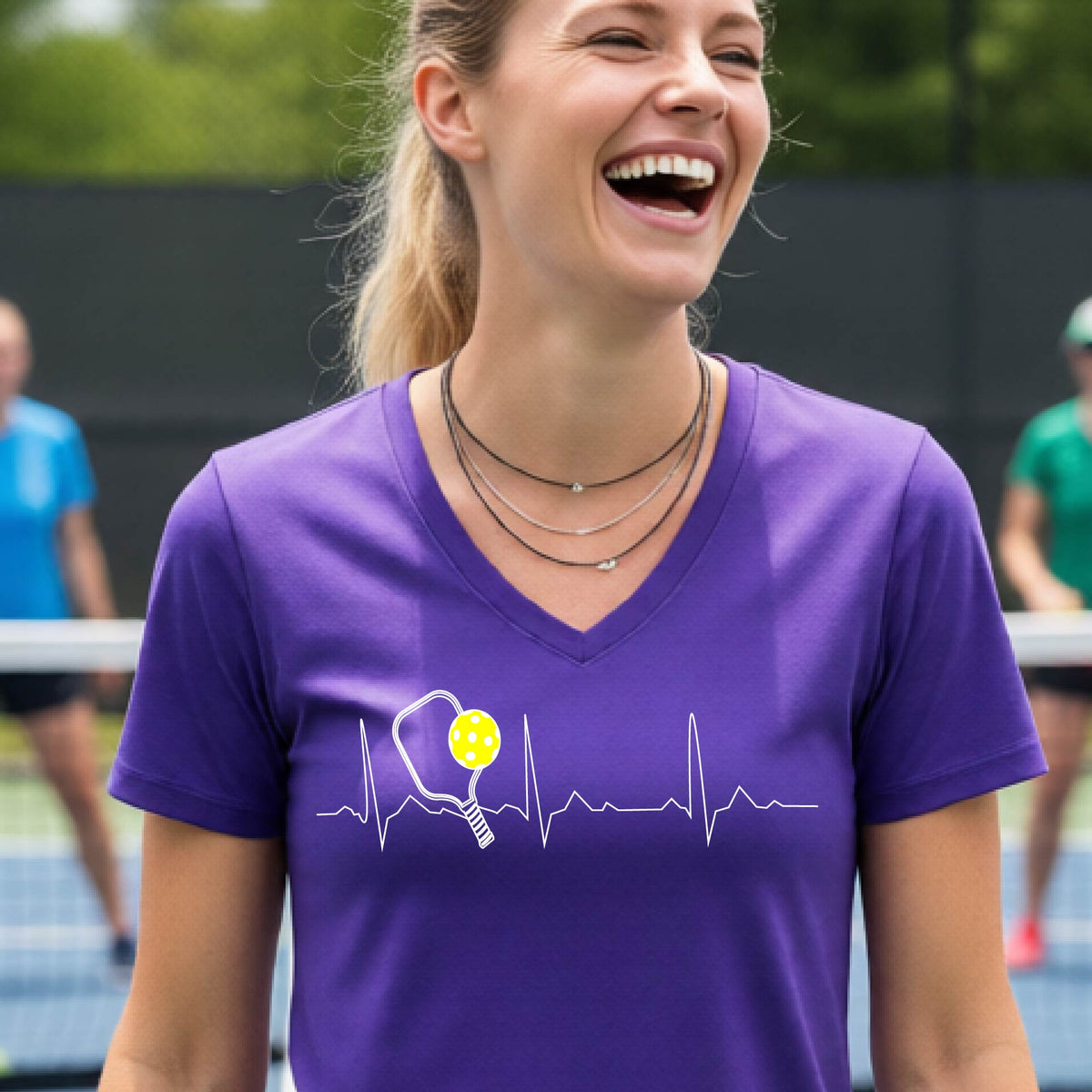 A close-up of a womens in a purple short-sleeve v-neck t-shirt sporting a white pickleball graphic of a heartbeat EKG including a pickleball paddle and a yellow pickleball across the check, in front of a pickleball court.