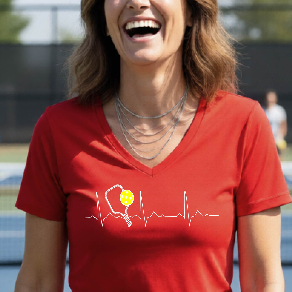 A close-up of a womens in a red short-sleeve v-neck t-shirt sporting a white pickleball graphic of a heartbeat EKG including a pickleball paddle and a yellow pickleball across the check, in front of a pickleball court.