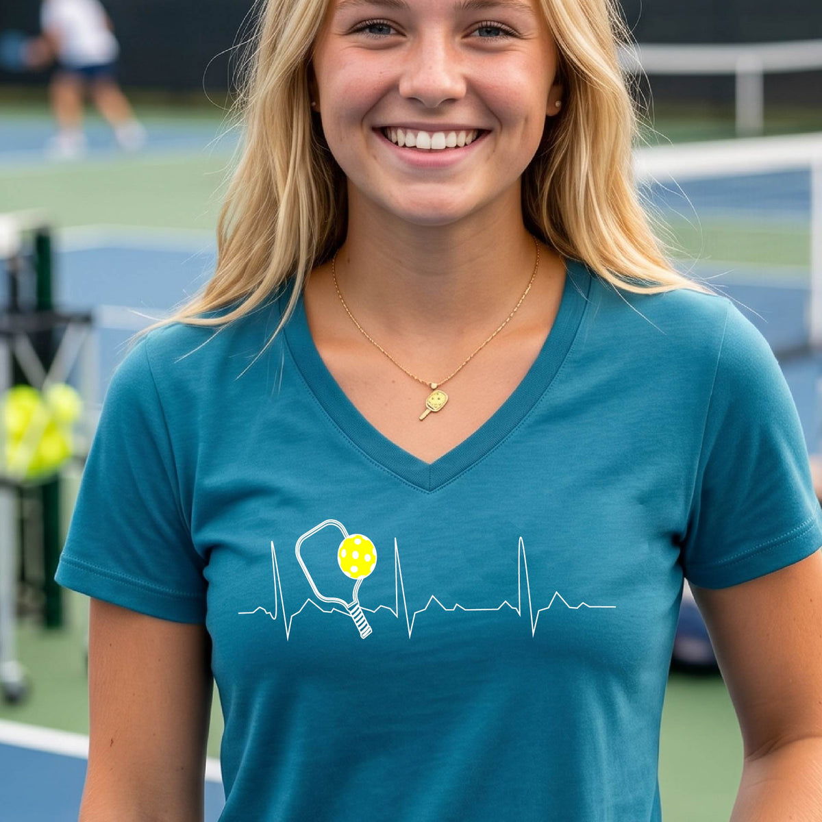 A close-up of a womens in a teal short-sleeve v-neck t-shirt sporting a white pickleball graphic of a heartbeat EKG including a pickleball paddle and a yellow pickleball across the check, in front of a pickleball court.