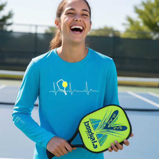 A close-up of a young person wearing bright blue long-sleeve t-shirt showing a white pickleball graphic of a heartbeat EKG including a pickleball paddle and a yellow pickleball, in front of a pickleball court.