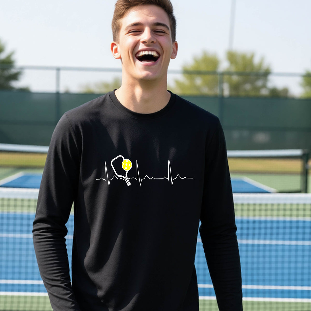 A close-up of a young person wearing black long-sleeve t-shirt showing a white pickleball graphic of a heartbeat EKG including a pickleball paddle and a yellow pickleball, in front of a pickleball court.