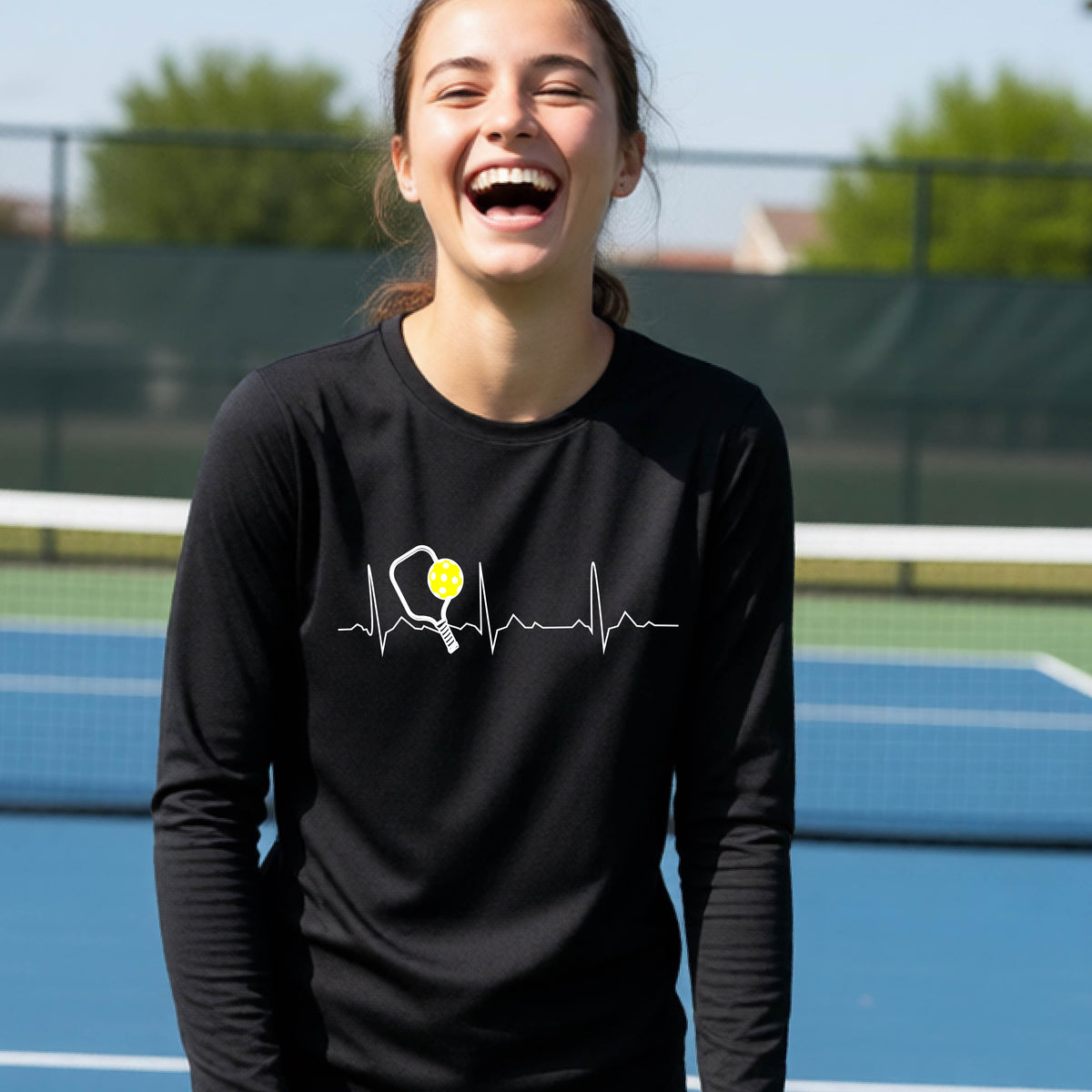 A close-up of a young person wearing black long-sleeve t-shirt showing a white pickleball graphic of a heartbeat EKG including a pickleball paddle and a yellow pickleball, in front of a pickleball court.