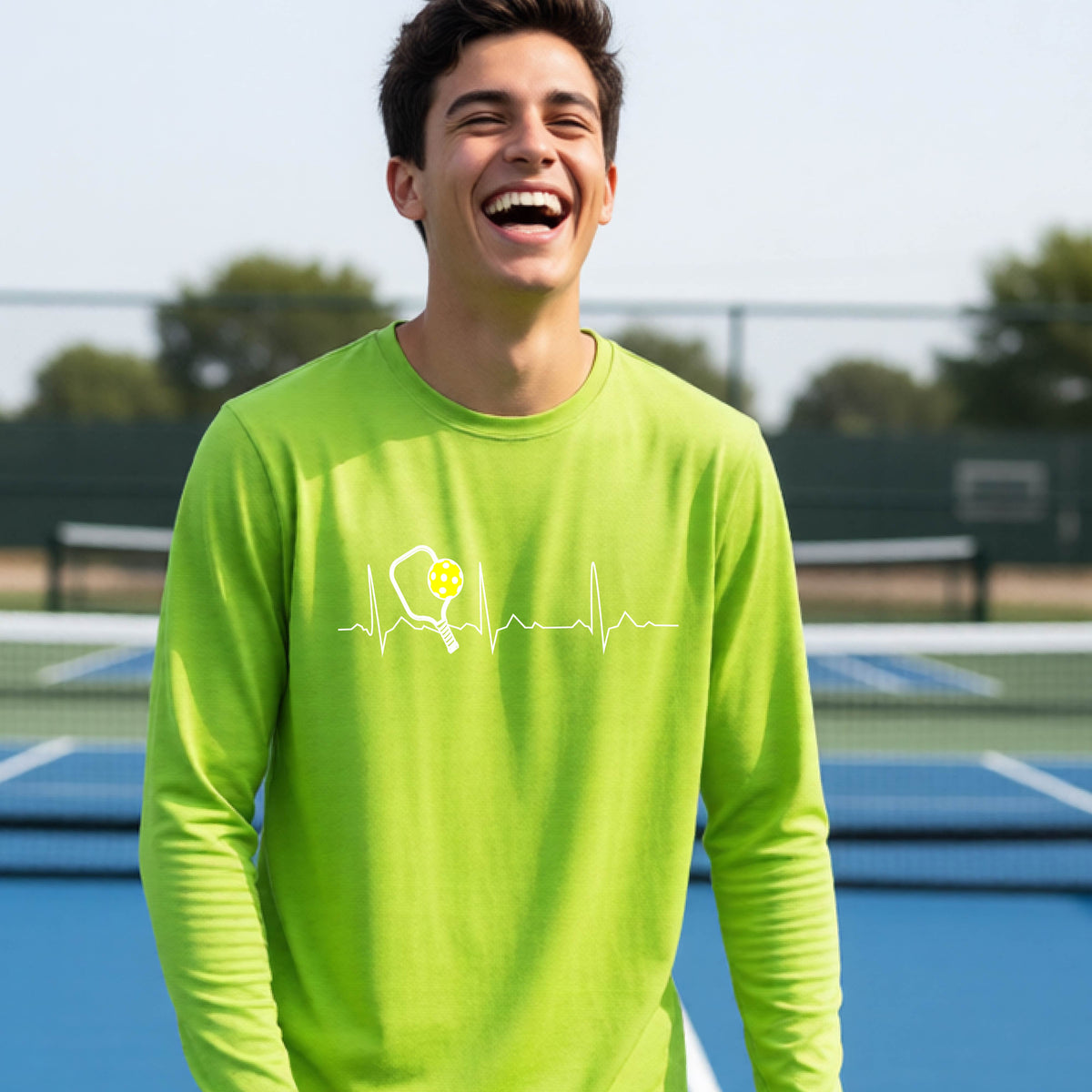 A close-up of a young person wearing lime green long-sleeve t-shirt showing a white pickleball graphic of a heartbeat EKG including a pickleball paddle and a yellow pickleball, in front of a pickleball court.