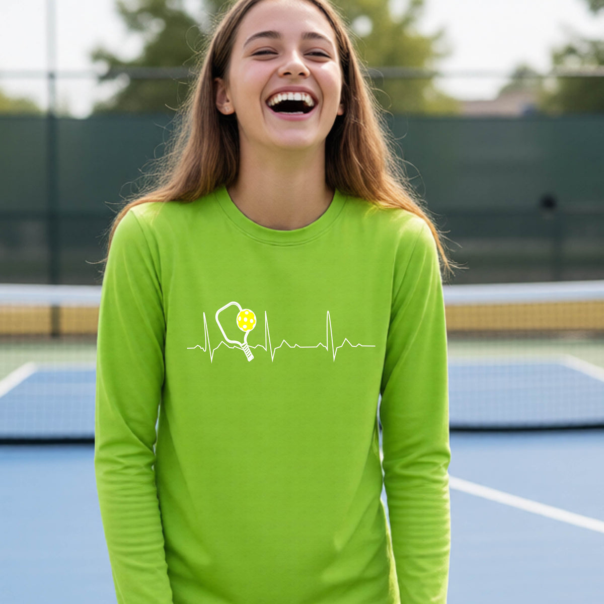 A close-up of a young person wearing lime green long-sleeve t-shirt showing a white pickleball graphic of a heartbeat EKG including a pickleball paddle and a yellow pickleball, in front of a pickleball court.
